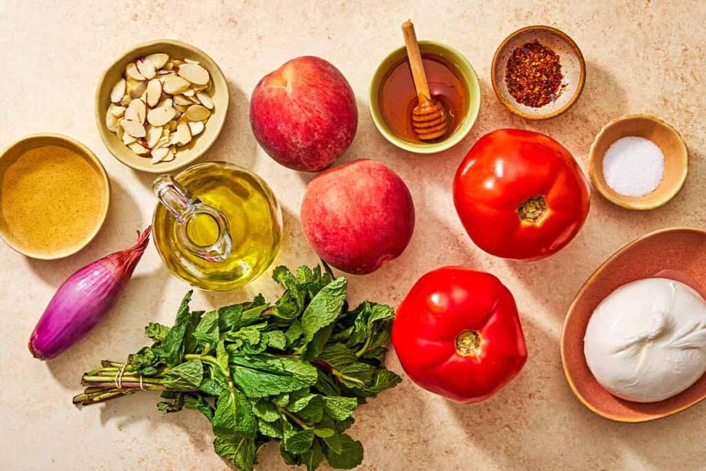 Ingredients for peach burrata salad including peaches, tomatoes, burrata, sliced almonds, mint, white wine vinegar, honey, salt, aleppo pepper, olive oil and a shallot.