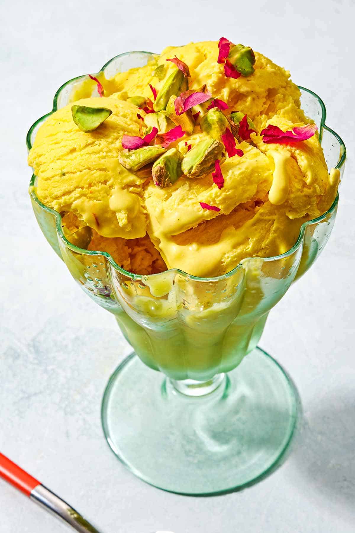 Persian ice cream (Bastani Sonnati) in a dessert goblet on a white surface. The ice cream is topped with rose petals an saffron.