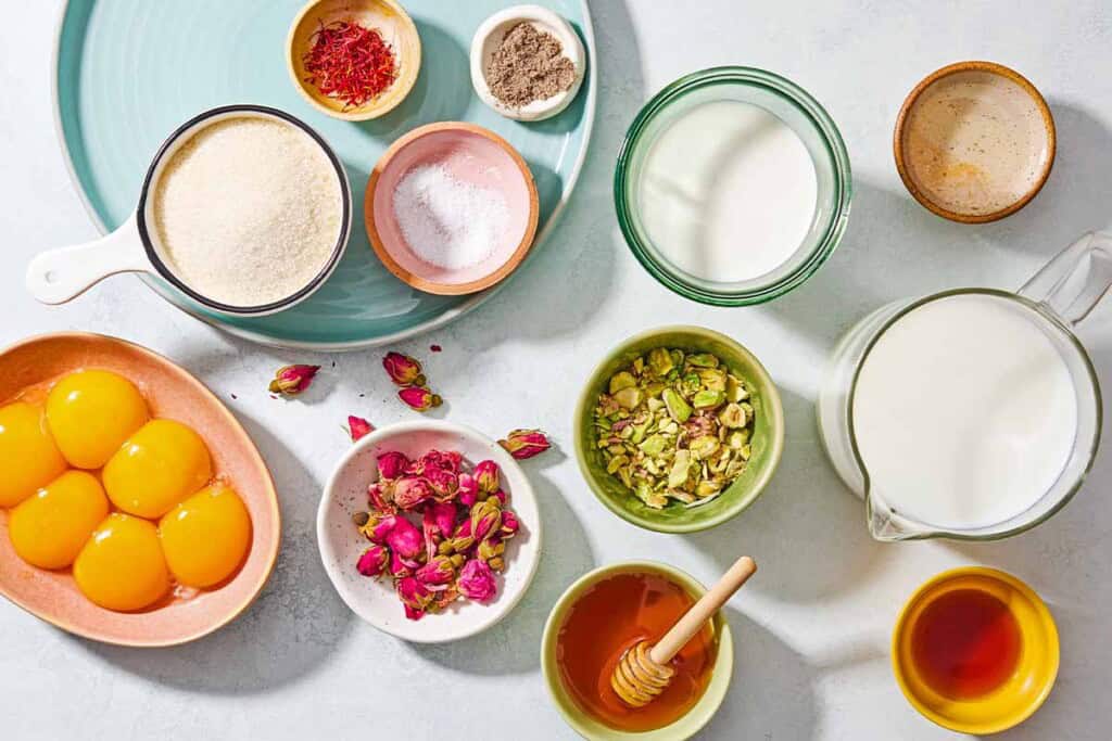 Ingredients for persian ice cream including egg yolks, sugar, milk, saffron, salt, rose water, vanilla extract, ground cardamom, cream, pistachios, dried rosebuds, and honey.