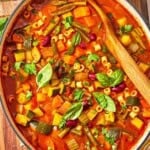Minestrone soup in a pot with a wooden spoon. Next to this is a bottle of olive oil and a bowl of grated parmesan cheese.
