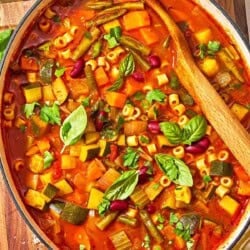 Minestrone soup in a pot with a wooden spoon. Next to this is a bottle of olive oil and a bowl of grated parmesan cheese.