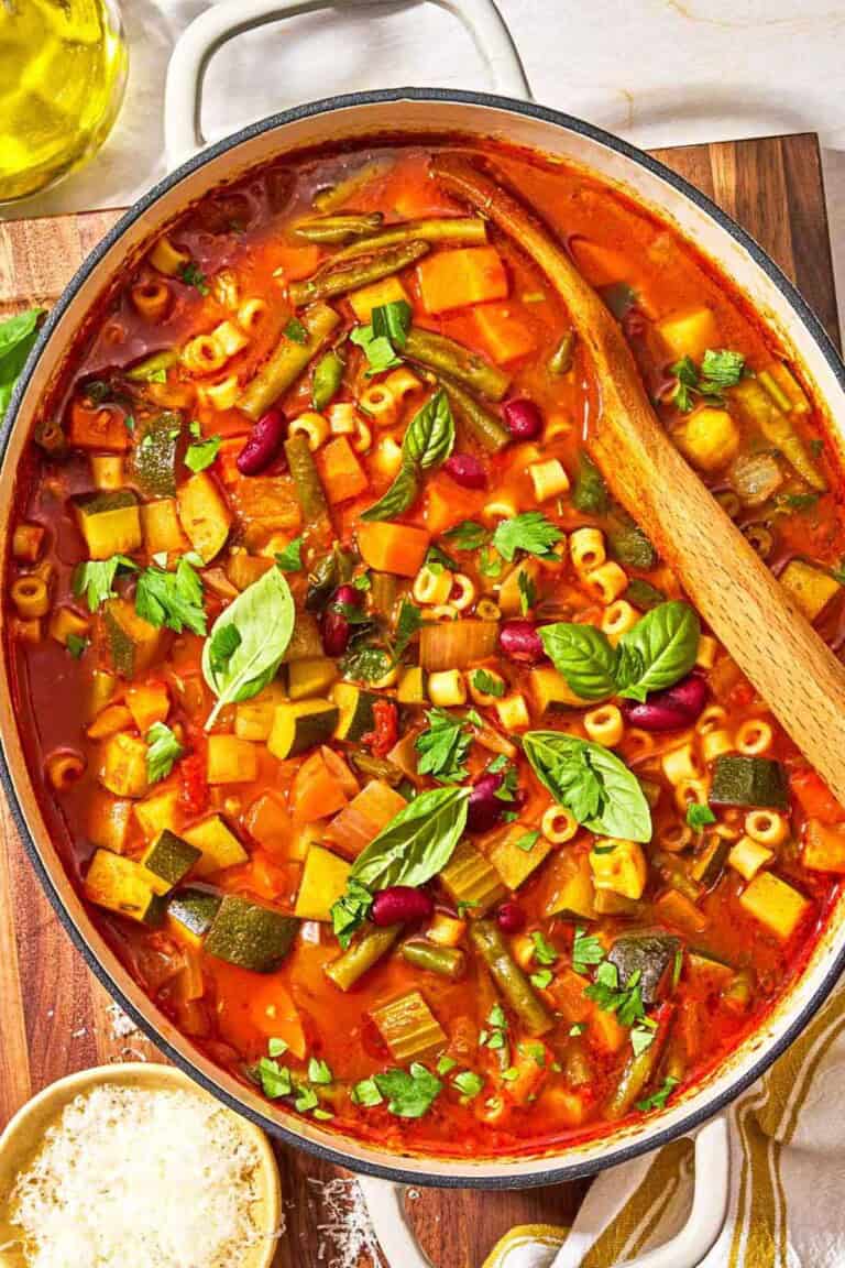 Minestrone soup in a pot with a wooden spoon. Next to this is a bottle of olive oil and a bowl of grated parmesan cheese.