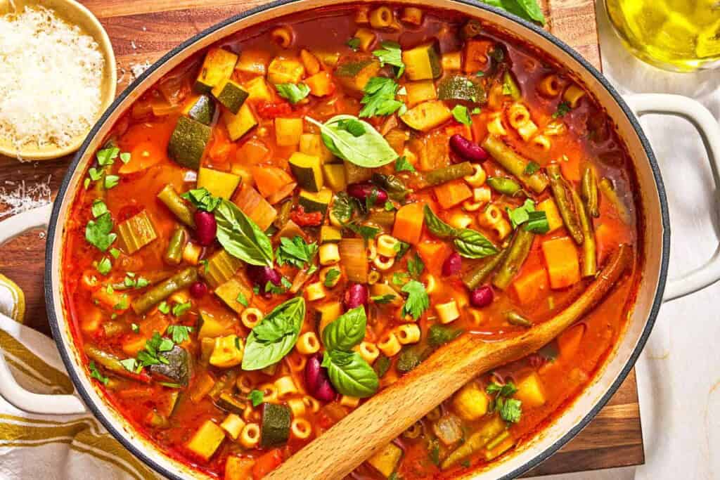 Minestrone soup in a pot with a wooden spoon. Next to this is a bottle of olive oil and a bowl of grated parmesan cheese.