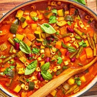 Minestrone soup in a pot with a wooden spoon. Next to this is a bottle of olive oil and a bowl of grated parmesan cheese.