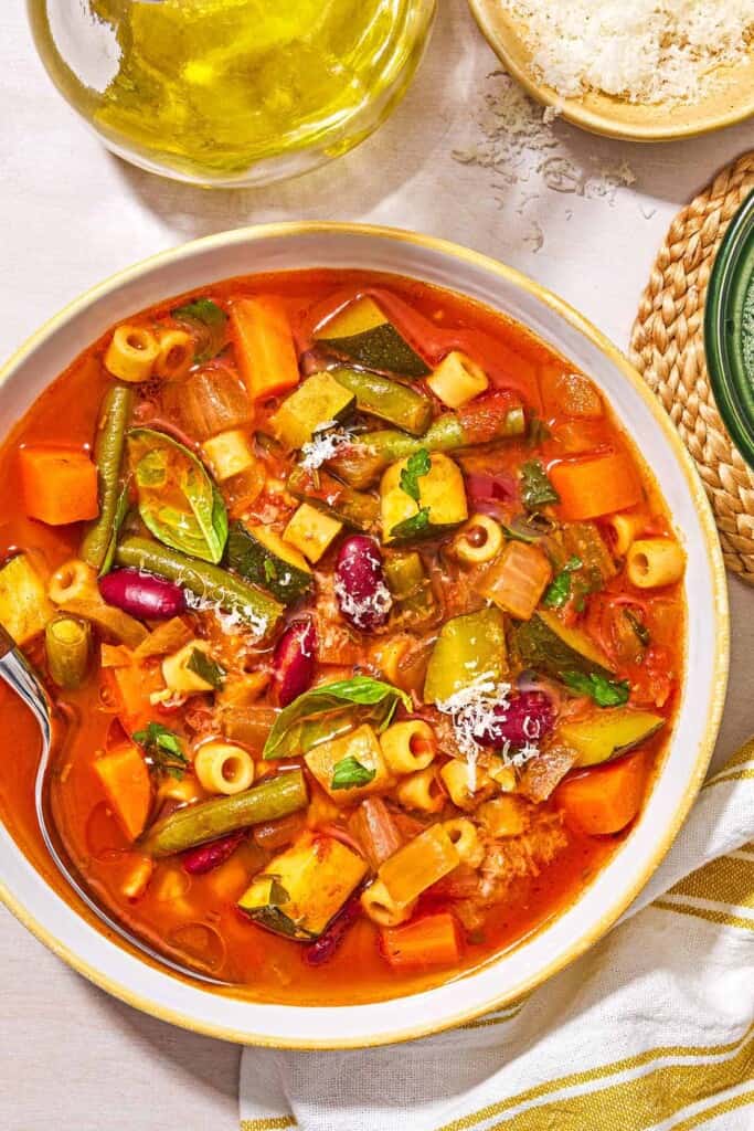 Minestrone soup in a bowl with a spoon next to a bottle of olive oil and a bowl of grated parmesan cheese.