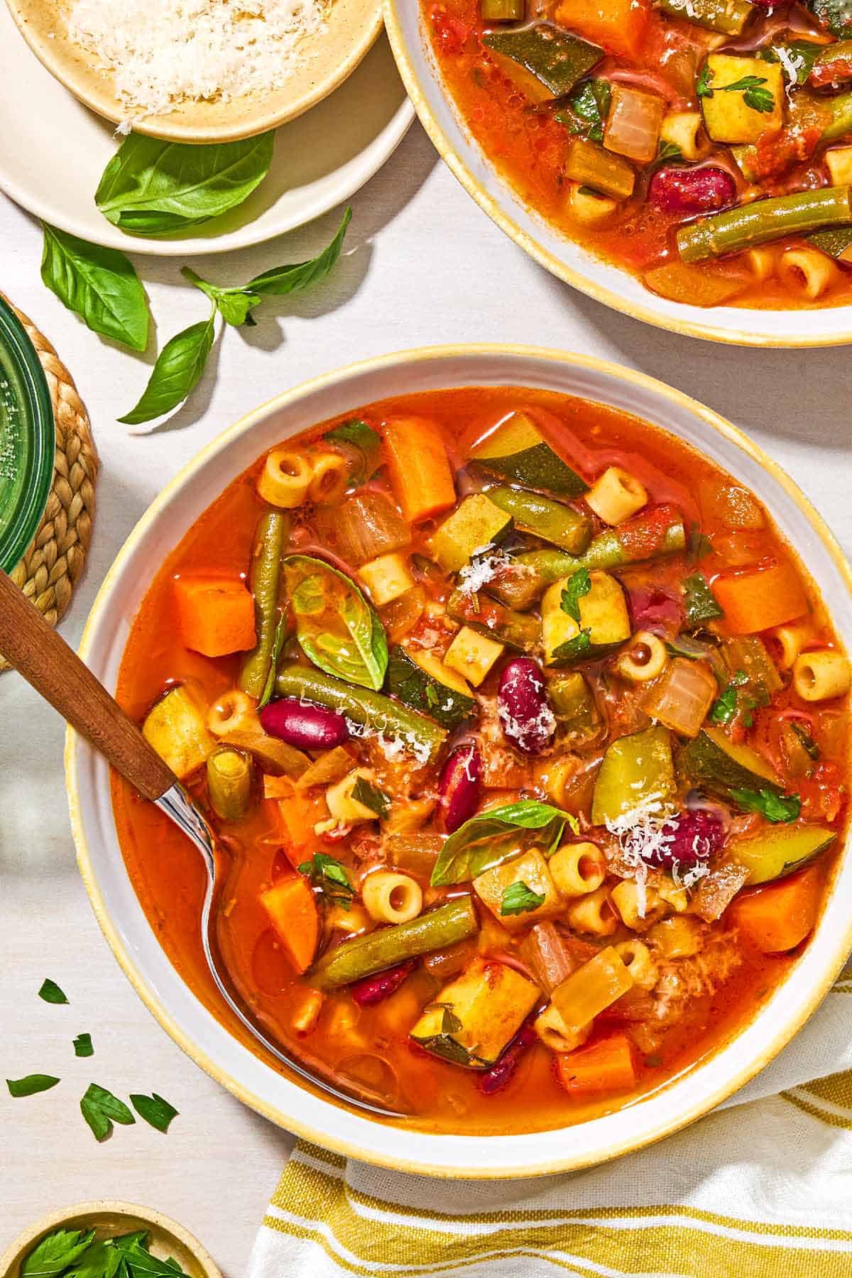 Minestrone soup in a bowl with a spoon next to another bowl of the soup, a bowl of grated parmesan cheese, and a kitchen towel.