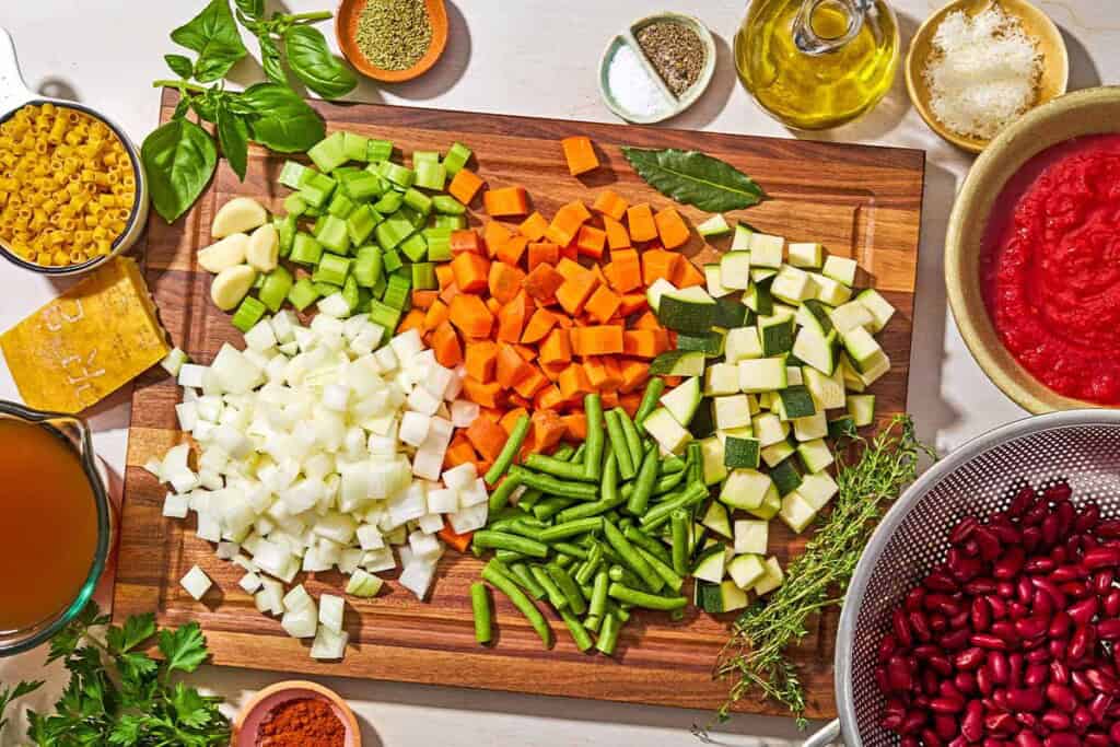 The ingredients for minestrone soup including ditalini pasta, chopped onion, carrots, celery, and zucchini, green beans, garlic, a bay leaf, olive oil, salt, black pepper, sweet paprika, rosemary, crushed tomatoes, broth, thyme, parmesan cheese rind, kidney beans, parsley, basil and grated parmesan cheese.