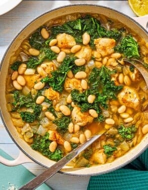 White bean and kale soup in a pot with a ladle next to a bottle oil and a lemon half.