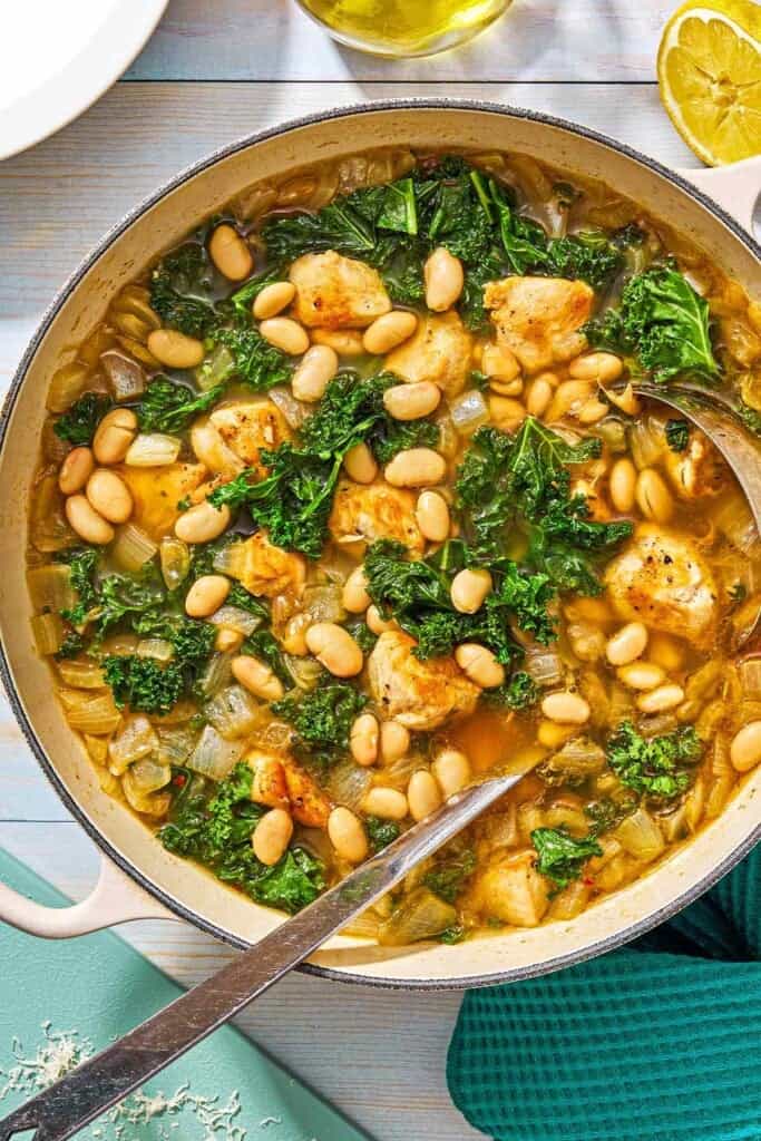 White bean and kale soup in a pot with a ladle next to a bottle oil and a lemon half.