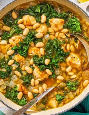 White bean and kale soup in a pot with a ladle next to a kitchen towel and 2 lemon halves.