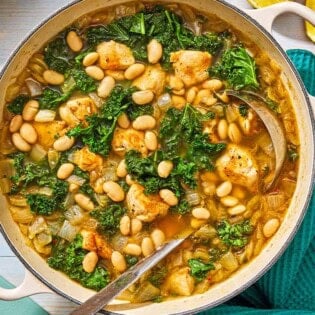 White bean and kale soup in a pot with a ladle next to a kitchen towel and 2 lemon halves.