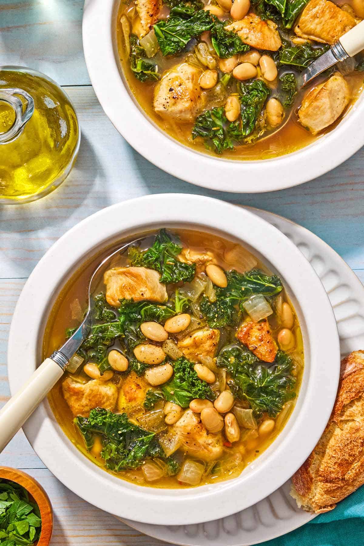 Two bowls of white bean and kale soup with spoon, one resting on a plate with a piece of crusty bread. Next to these is a bottle of olive oil and a small bowl of chopped parsley.