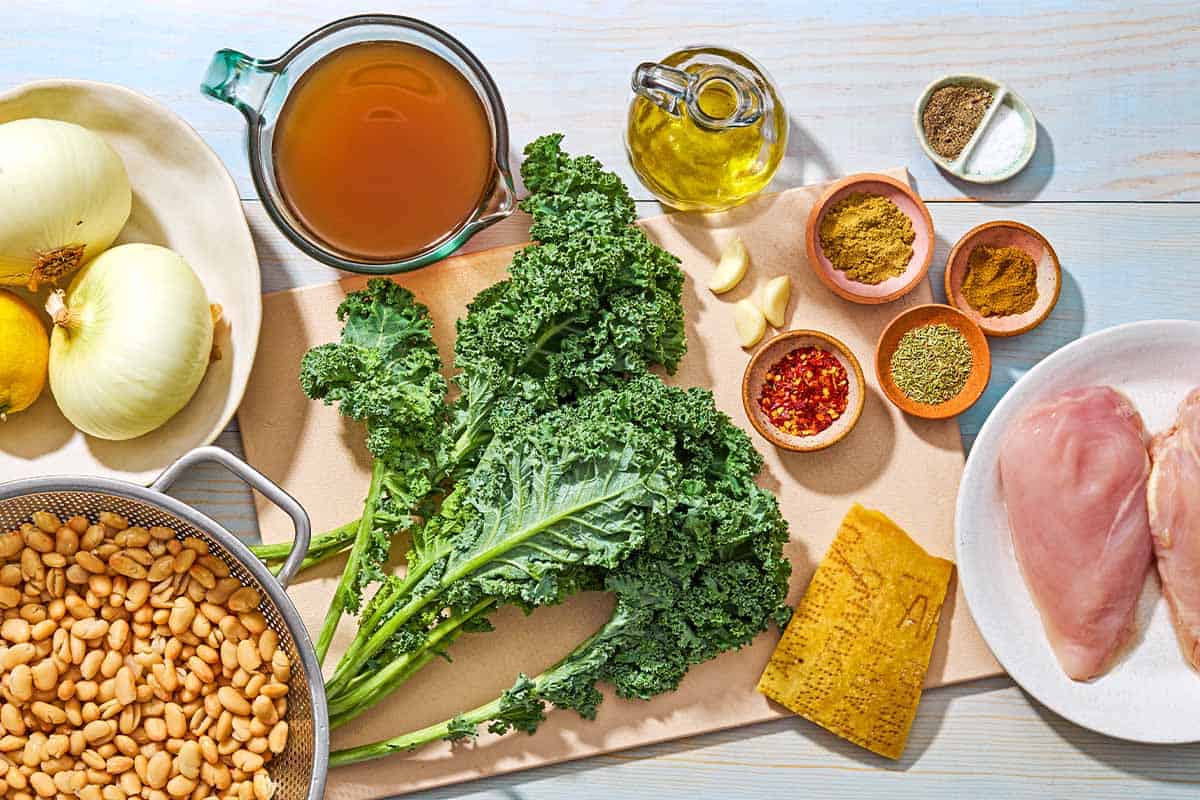 Ingredients for white bean and kale soup including chicken, olive oil, salt, black pepper, onions, garlic, red pepper flakes, ground coriander, ground cumin, dried rosemary, white beans, broth, kale, a parmesan rind and lemon.