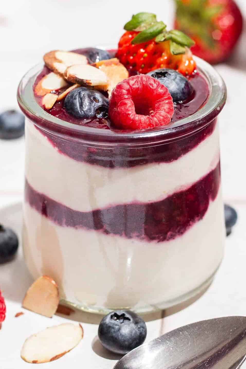 A close up of a yogurt parfait in a glass jar topped with berries. Next to this is a spoon and various berries scattered about.