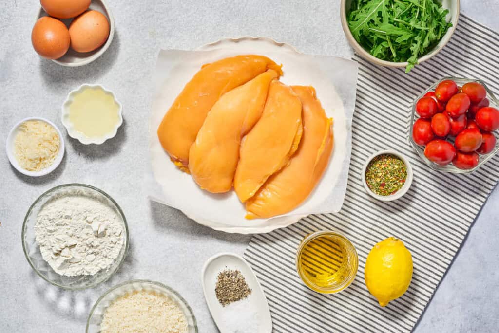 Ingredients for chicken milanese including chicken breasts, eggs, salt, pepper, flour, breadcrumbs, Parmigiano-Reggiano cheese, italian seasoning, olive oil, arugula, lemon, and cherry tomatoes.