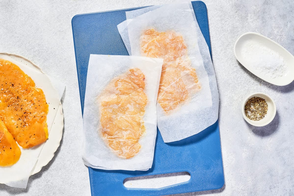 2 flattened chicken cutlets in between piece of parchment paper on a cutting board. Next to this is a plate with 2 seasoned, uncooked cutlets, and bowls of salt and black pepper.