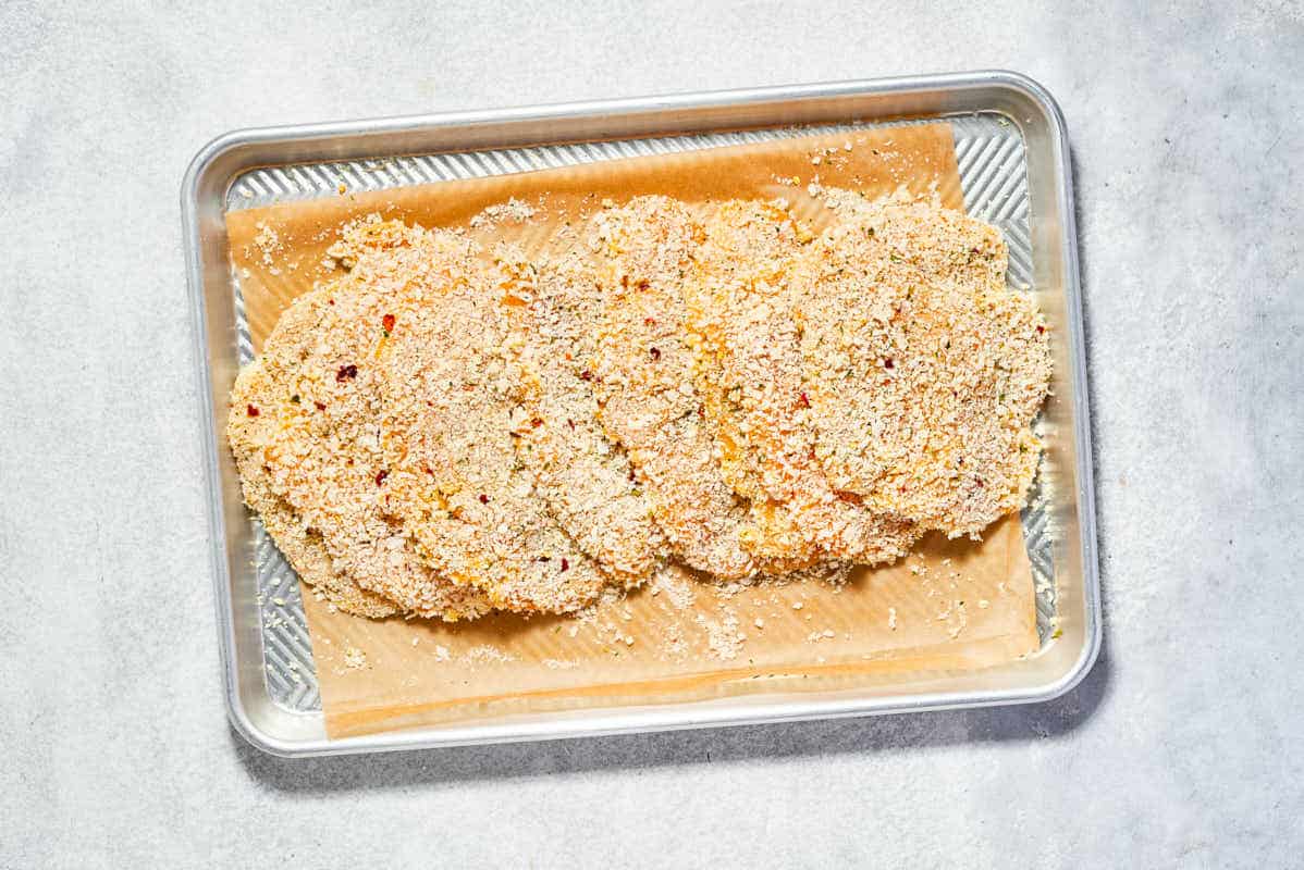 Several breaded but uncooked chicken cutlets on a parchment lined sheet pan.