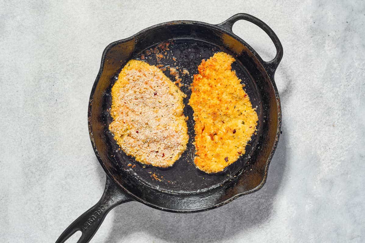 Two breaded chicken cutlets being fried in a cast iron skillet.