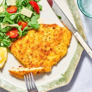 A piece of the chicken milanese on a plate with a serving of the arugula salad and a knife and fork. Next to this is a glass of sparkling water.