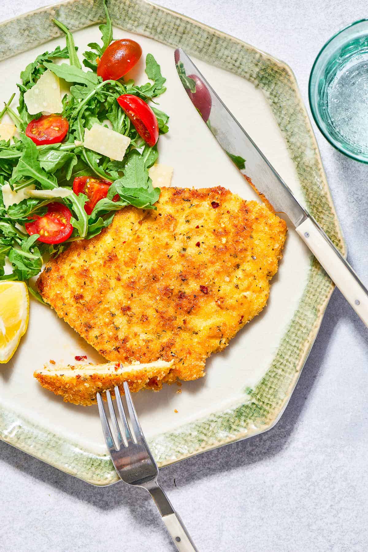 A piece of the chicken milanese on a plate with a serving of the arugula salad and a knife and fork. Next to this is a glass of sparkling water.
