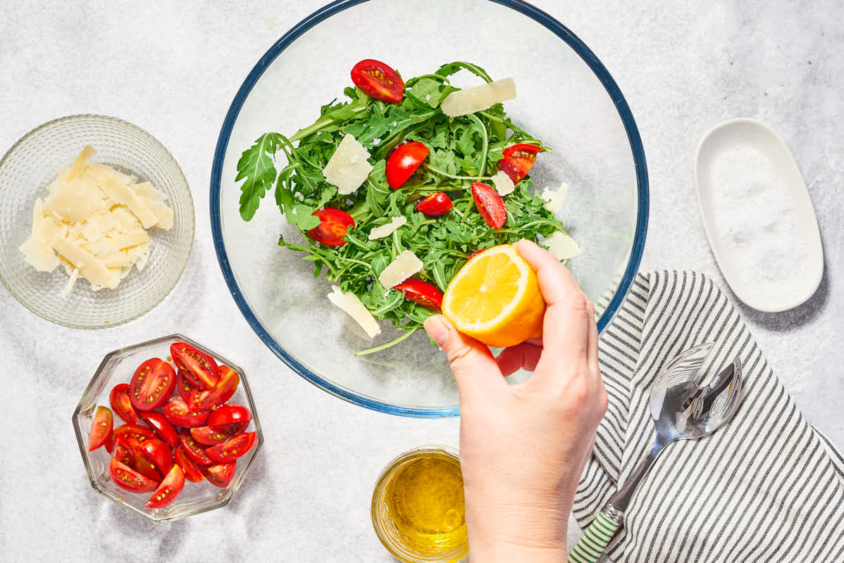 A lemon half being squeezed of the arugula salad in a mixing bowl. Next to this is a kitchen towel, and bowls of shaved parmigiano reggiano cheese, sliced cherry tomatoes, and salt.