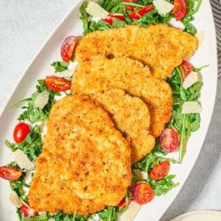 Chicken milanese cutlets on a bed of arugula salad on a serving platter. Next to this is a plate with 2 forks.