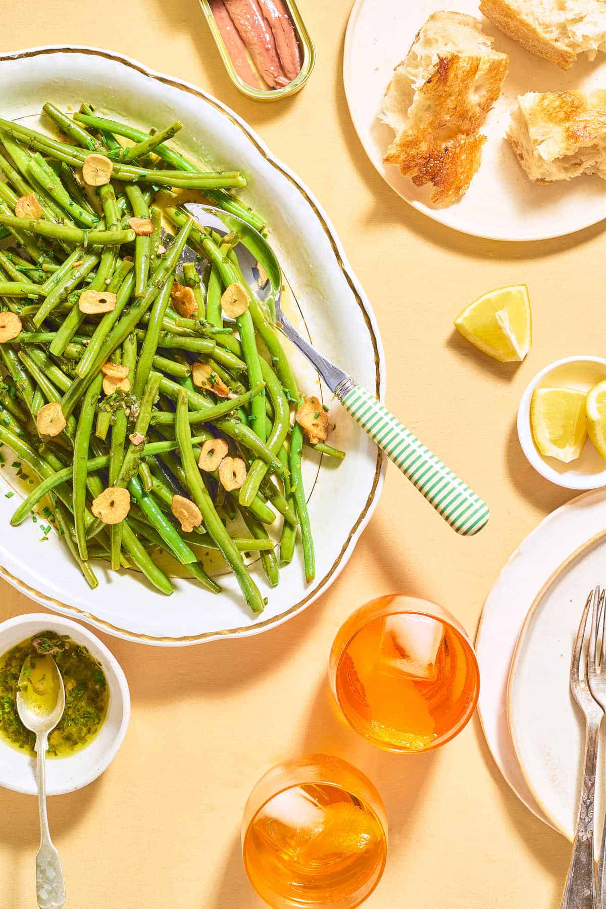 Italian green bean salad in a serving bowl with a spoon next to a tin of anchovies, a plate of crusty bread pieces, a bowl of lemon wedges, a stack of 2 plates with 2 forks, 2 beverages, and a bowl of dressing with a spoon.