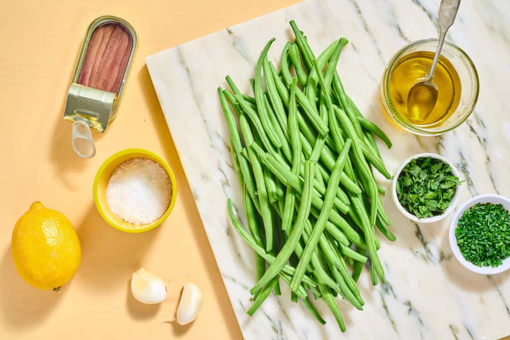 Ingredients for italian green bean salad including fresh green beans, salt, olive oil, garlic, chives, parsley, anchovy fillets, and lemon.