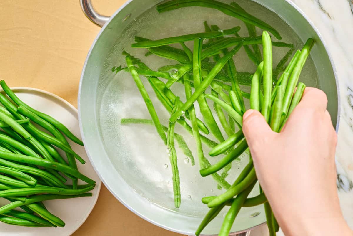Fresh green beans for the italian green bean salad being added to a pot of boiling water. Next to this is a plate with more fresh green beans.