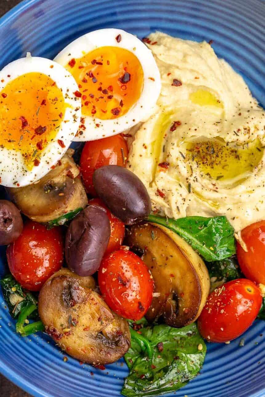A Mediterranean breakfast bowl with a soft-boiled egg, hummus and sauteed vegetables on a blue plate.