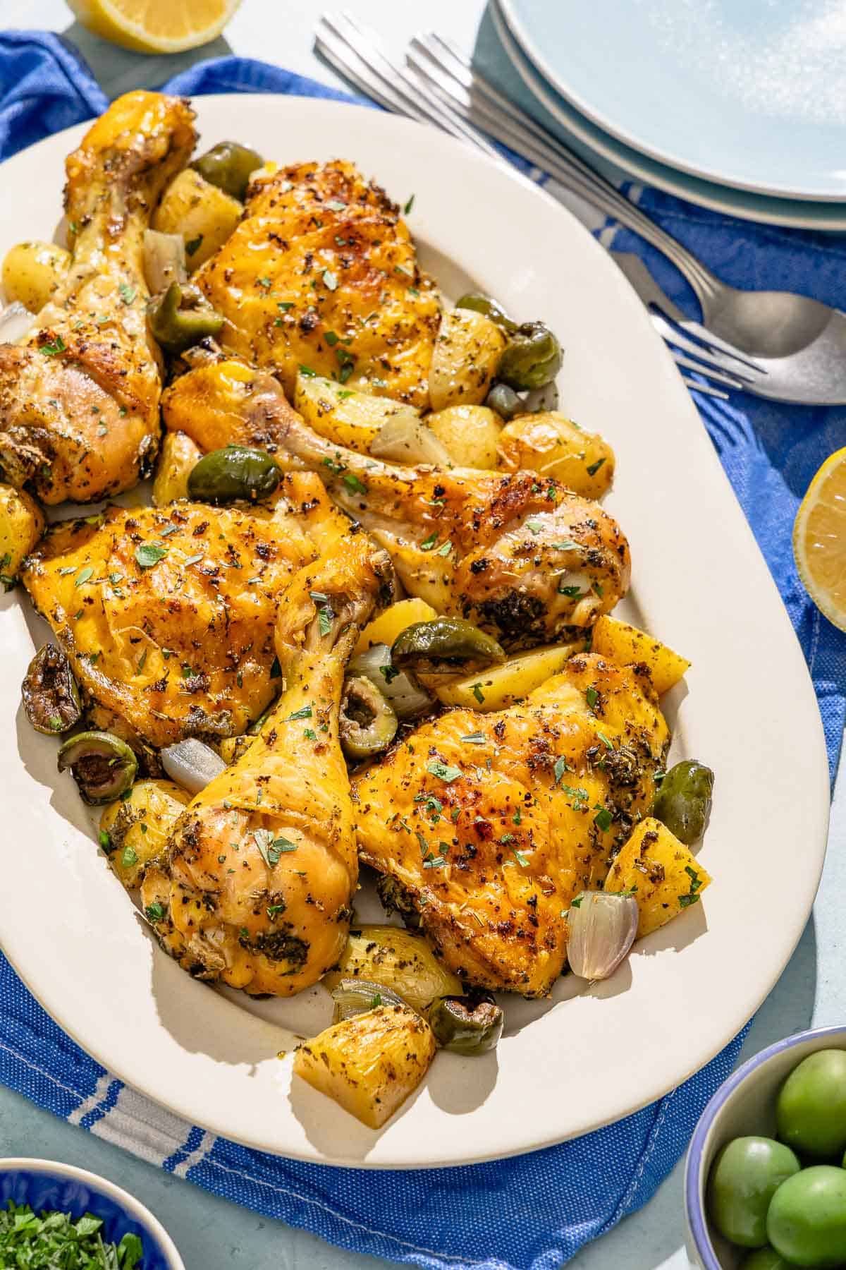 The chicken and potatoes recipe on a platter. Next to this is a stack of 2 plates, a fork and spoon, bowls of olives and chopped parsley, and two lemon halves.