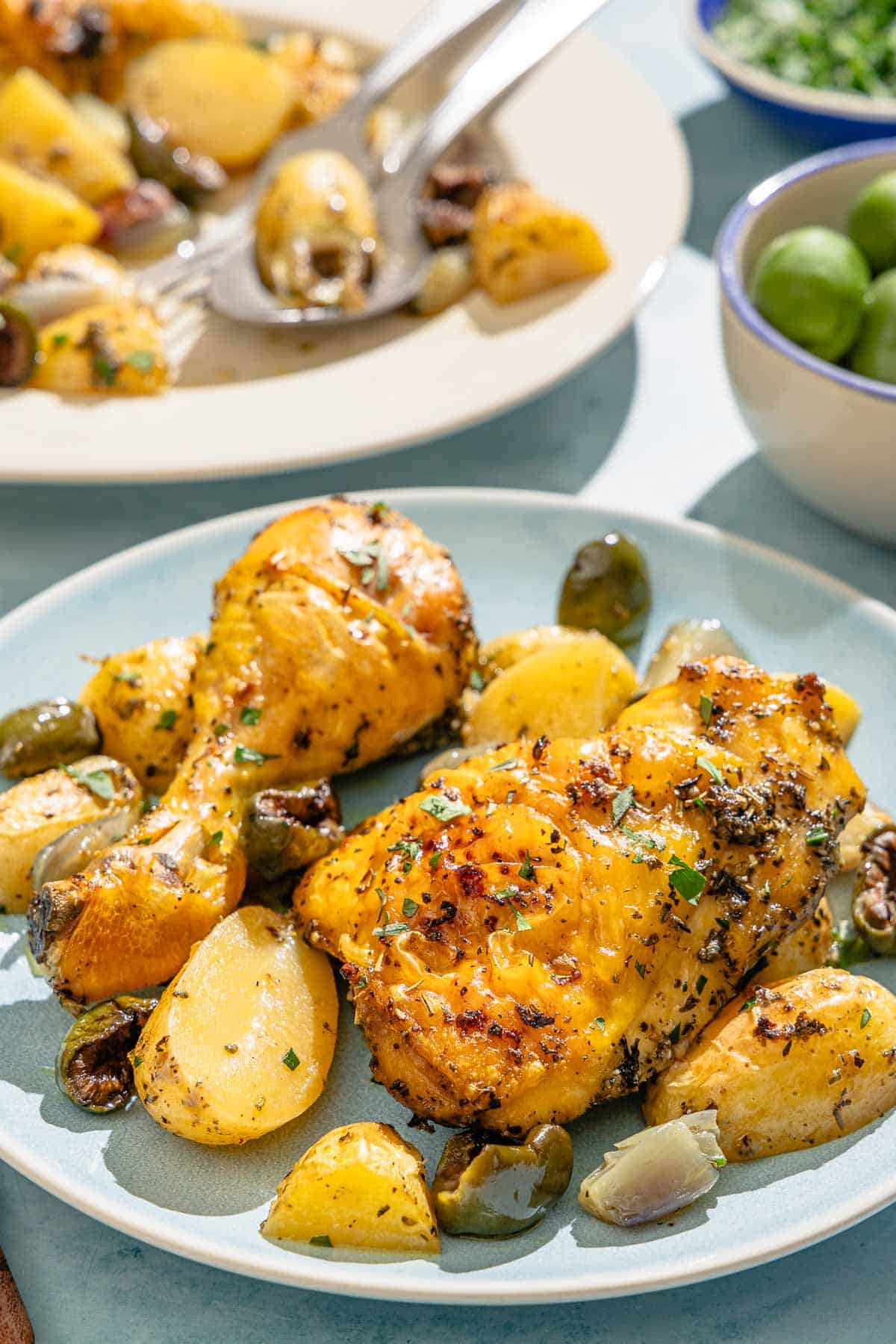 A serving of the chicken and potatoes on a plate. Next to this is a platter with the rest of the recipe with serving utensils and bowls of olives and chopped parsley.