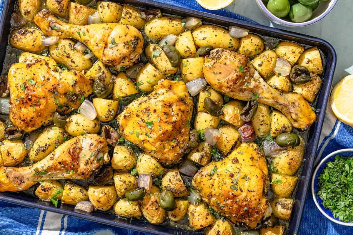 The baked chicken and potatoes recipe on a sheet pan. Next to this is a lemon slice and bowls of olives and chopped parsley.