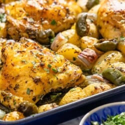 A close up of the baked chicken and potatoes recipe on a sheet pan next to a bowl of chopped parsley.