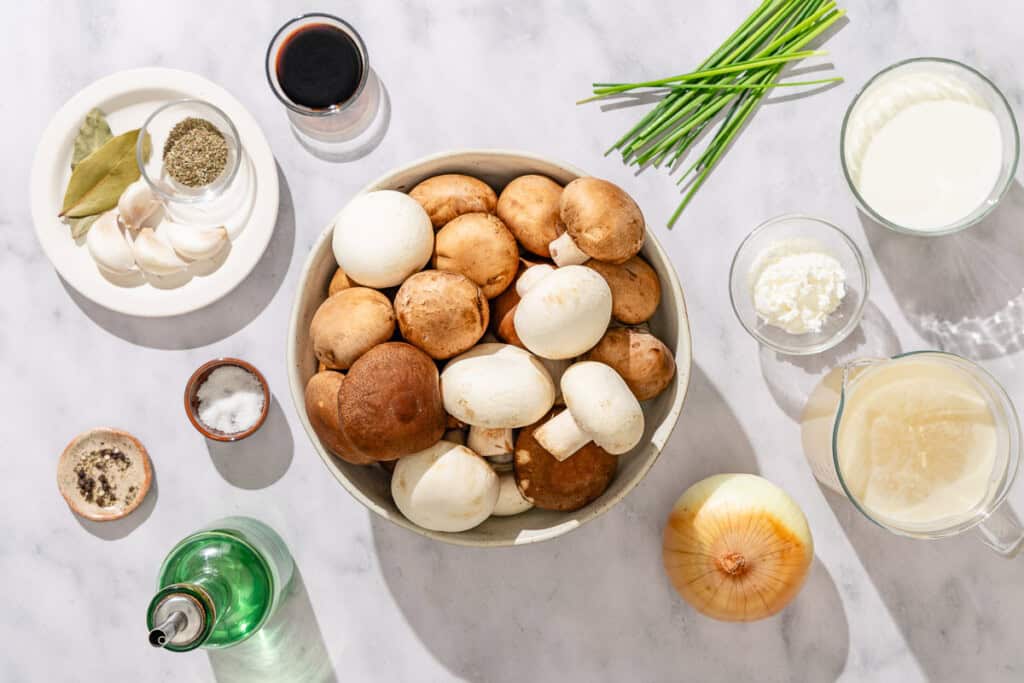 Ingredients for mushroom soup including mixed mushrooms, olive oil, onion, garlic, dried thyme, salt, black pepper, stock, bay leaves, milk, cornstarch, fresh chives.
