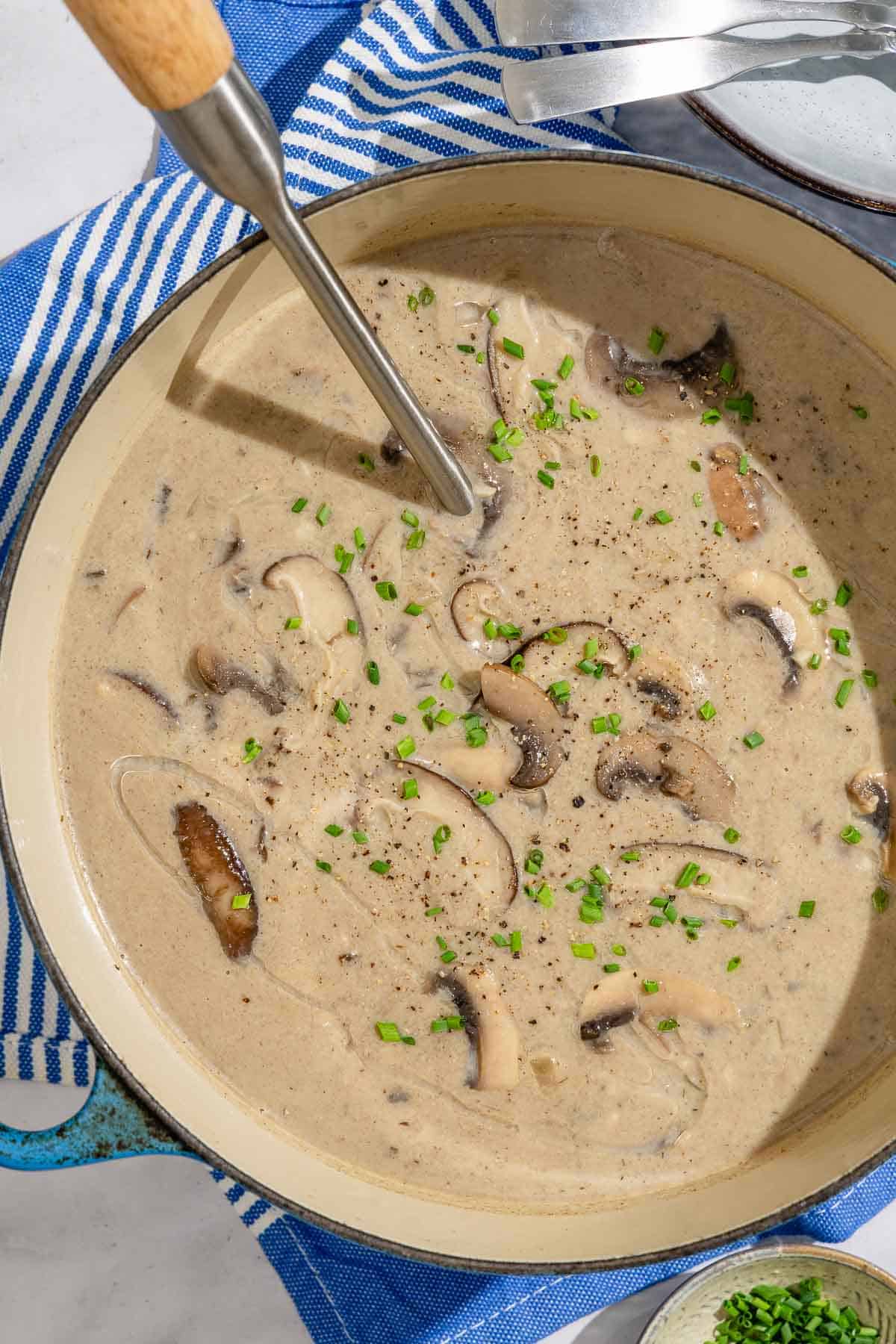 Mushroom soup in a large pot with a serving spoon. Next to this is a stack of bowls with 2 spoons, and a small bowl of chopped chives.