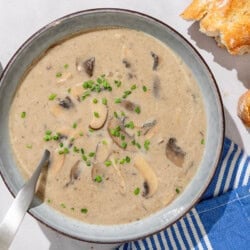 An overhead photo of mushroom soup in a bowl with a spoon. Next to this is a cloth napkin, a small bowl of chopped chives and some crusty bread.