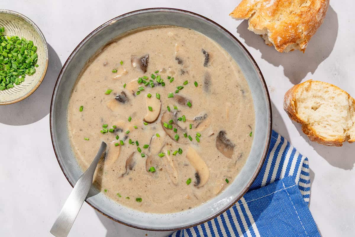 An overhead photo of mushroom soup in a bowl with a spoon. Next to this is a cloth napkin, a small bowl of chopped chives and some crusty bread.