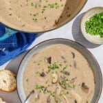 An overhead photo of mushroom soup in a bowl with a spoon. Next to this is the rest of the soup in a pot, a small bowl of chopped chives and some crusty bread.