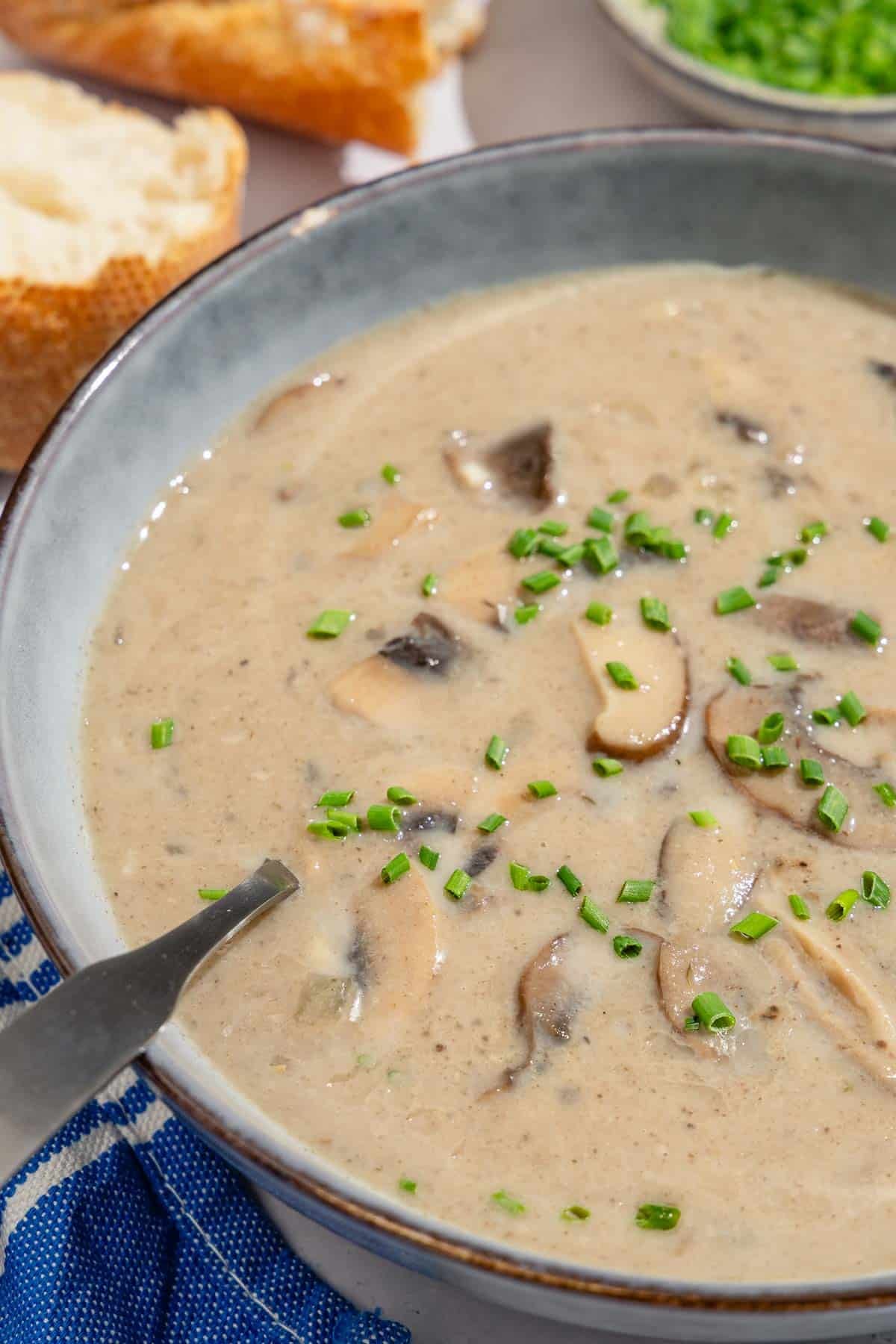 A close up of mushroom soup in a bowl with a spoon. Next to this is a small bowl of chopped chives and some crusty bread.