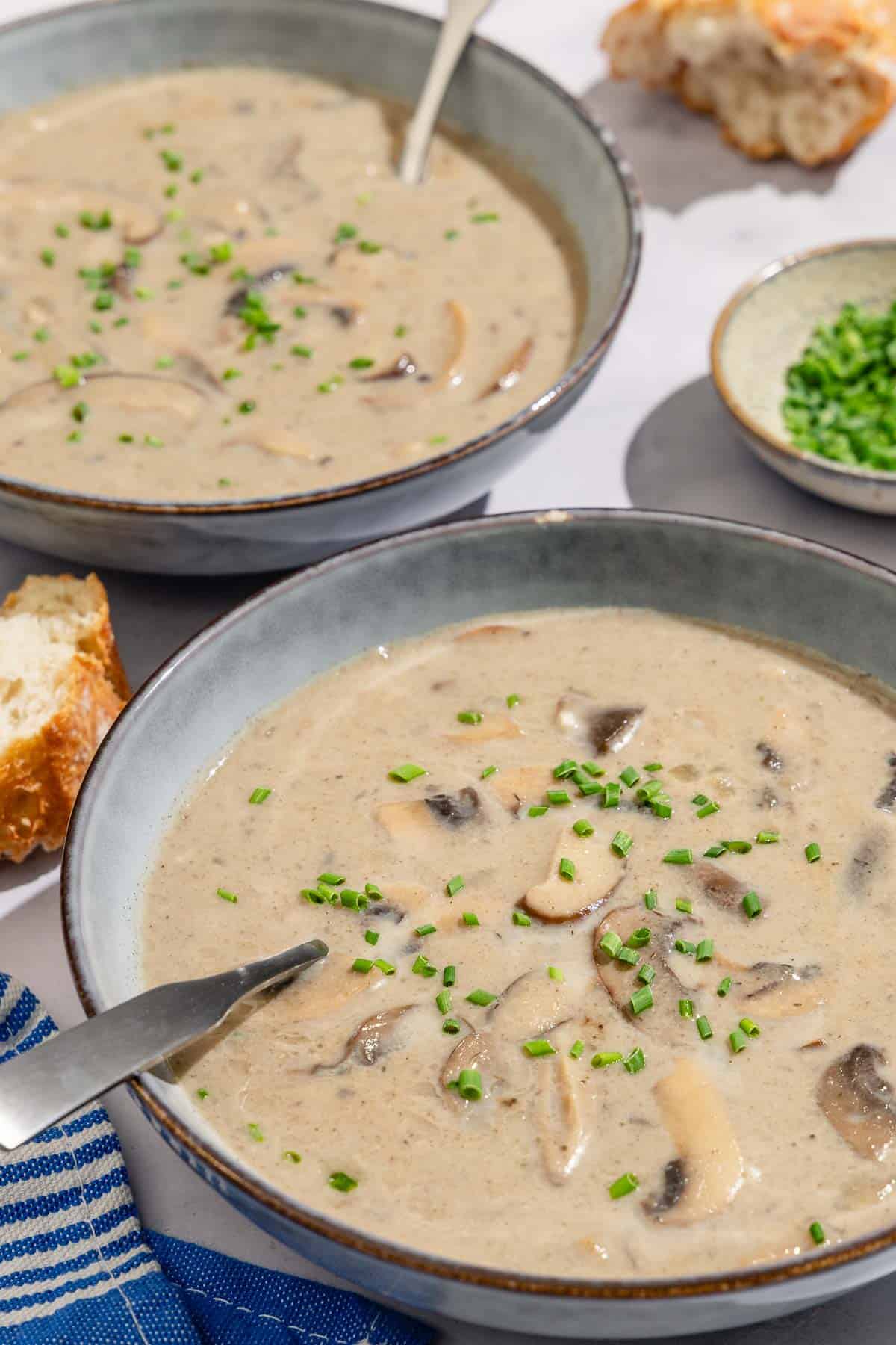 A close up of 2 bowls of mushroom soup with spoons. Next to these are pieces of crusty bread and a small bowl of chopped chives.
