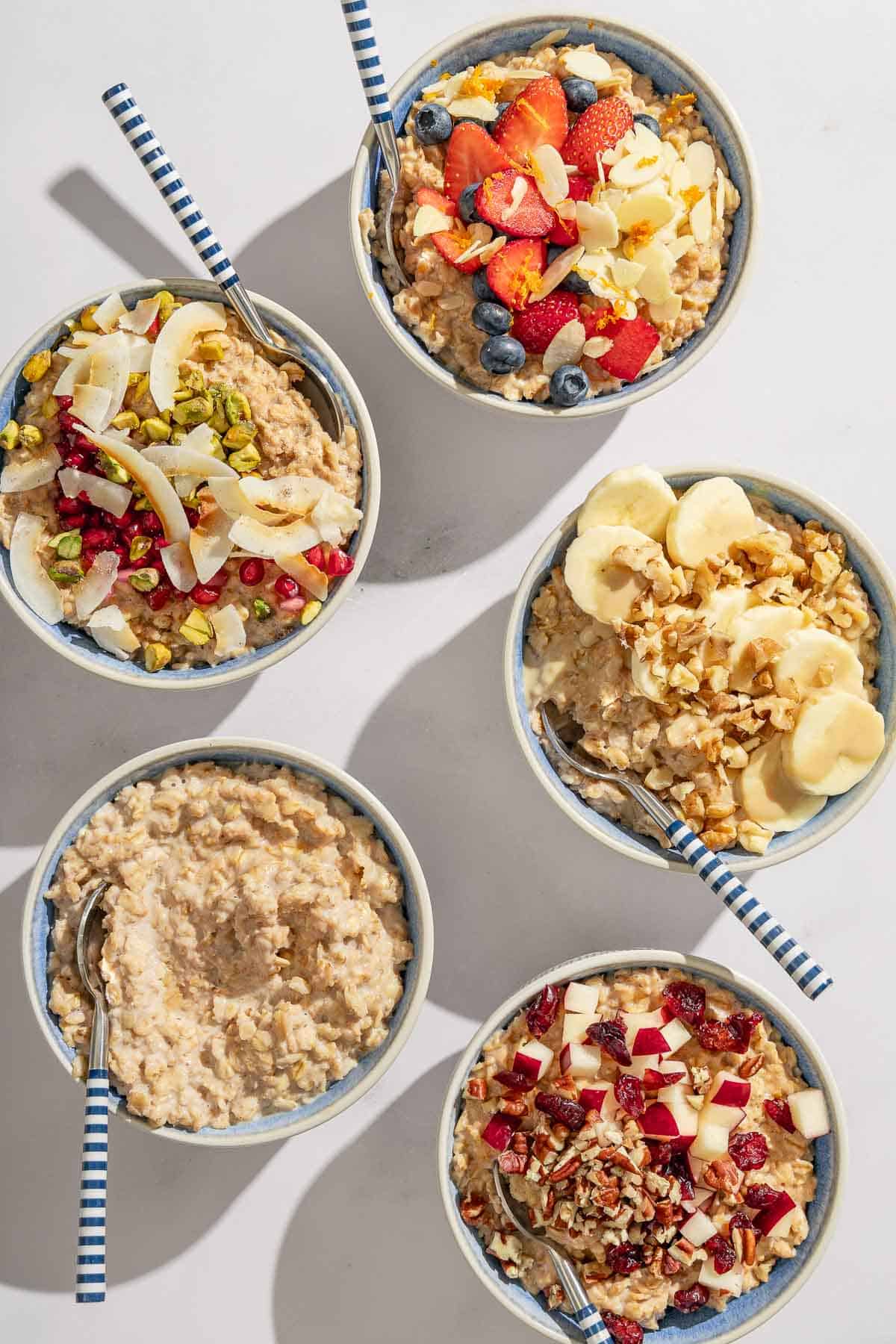 An overhead photo of 5 bowls of oatmeal with spoons. Four have various toppings and one is plain.
