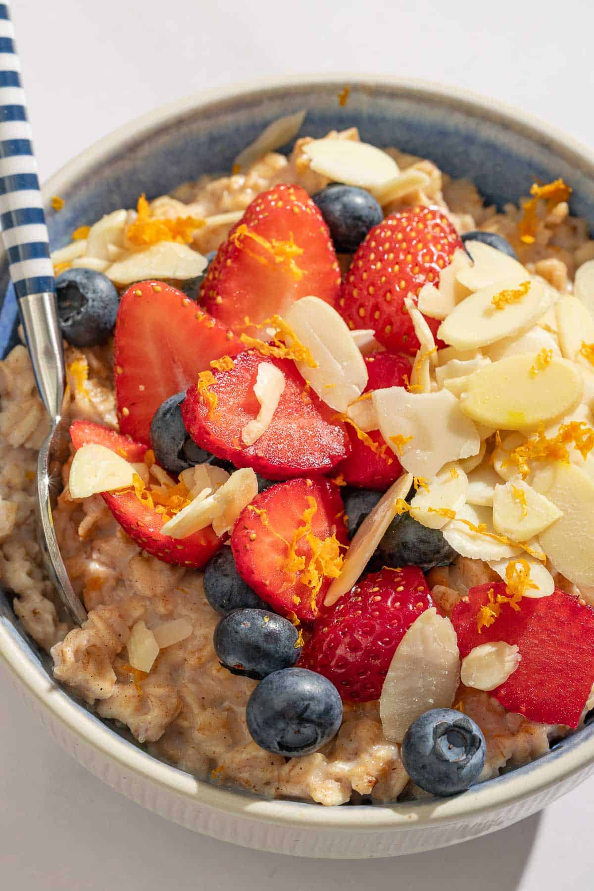 Oatmeal topped with strawberries, blueberries, chia seeds, almonds and orange zest in a bowl with a spoon.