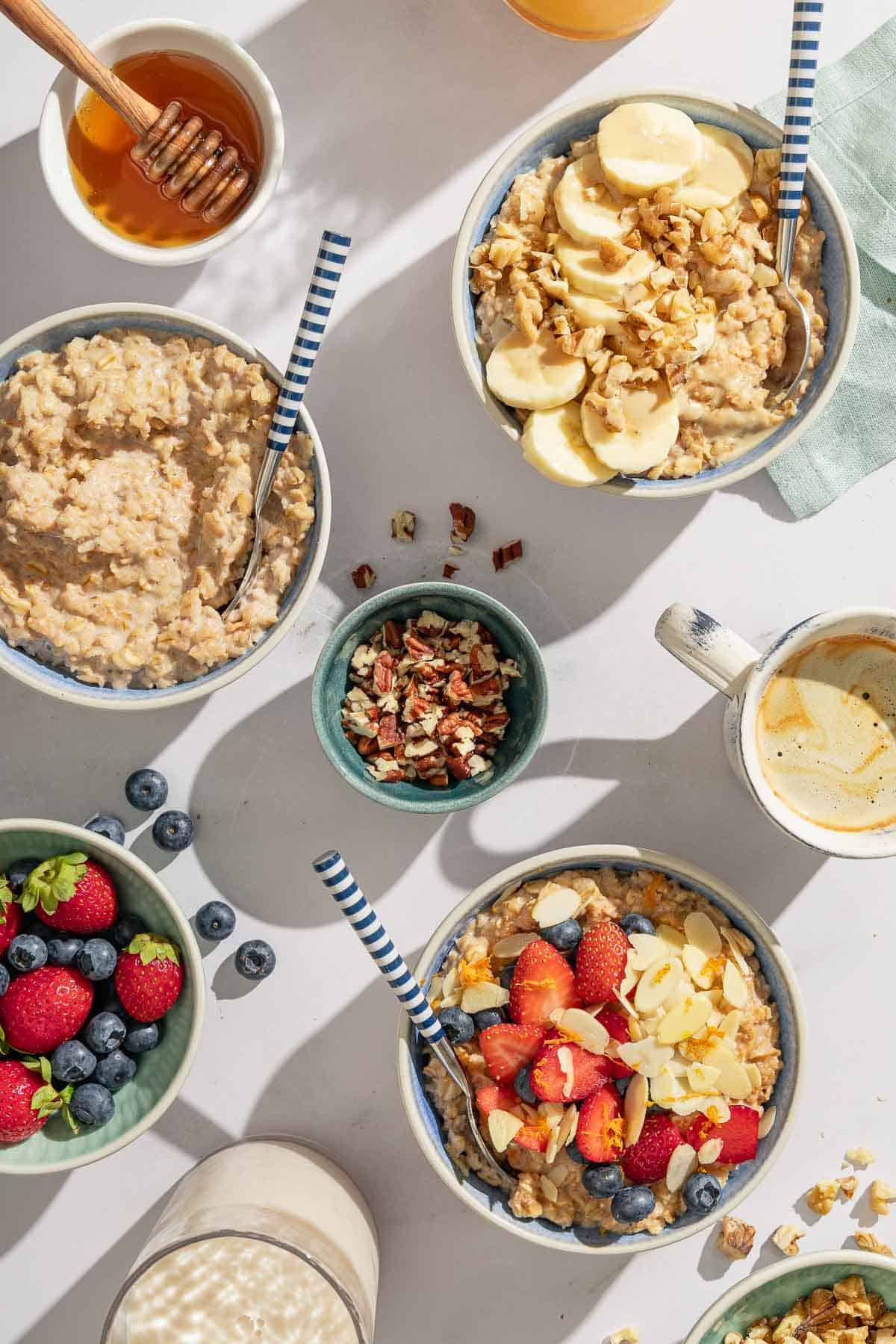 An overhead photo of three bowls of oatmeal with spoons. Two have various toppings and one is plain. next to these is a cup of coffee, a cup of milk, and bowls of nuts, berries and honey.