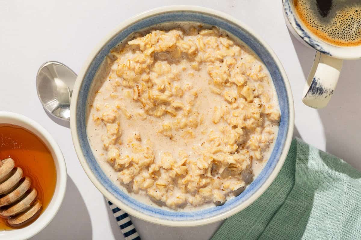 A bowl of oatmeal next to a spoon, a cup of coffee, a bowl of honey with a honey dipper and a cloth napkin.