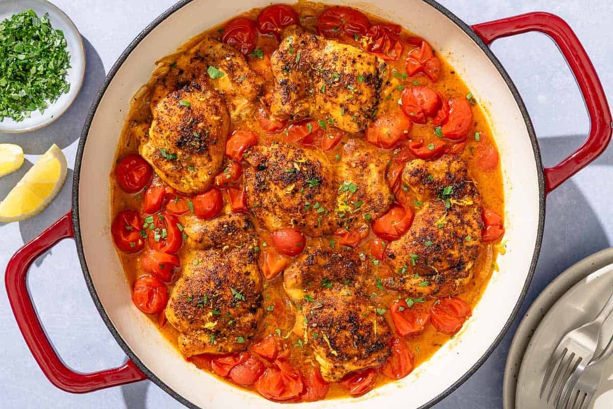 Chicken thighs in a cherry tomato sauce in a skillet next to a stack of 2 plates with forks, a small bowl of chopped parsley, and lemon wedges.