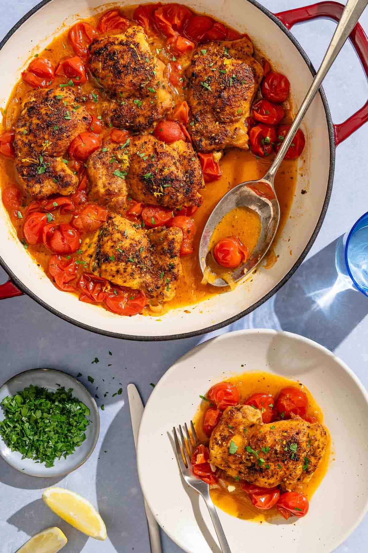 A serving of the skillet chicken thighs on a plate with a fork. Next to this is a knife, the rest of the recipe in a skillet with a serving spoon, a small bowl of chopped parsley, two lemon wedges and a glass of water.