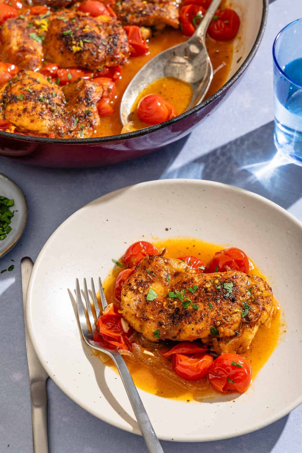 A serving of the skillet chicken thighs on a plate with a fork. Next to this is a knife, the rest of the recipe in a skillet with a serving spoon, and a glass of water.