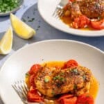 Two servings of the skillet chicken thighs on plates with forks. Next to these are 2 lemon wedges and a small bowl of chopped parsley.