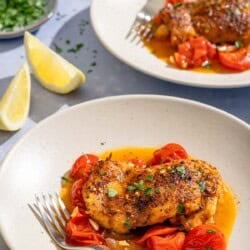 Two servings of the skillet chicken thighs on plates with forks. Next to these are 2 lemon wedges and a small bowl of chopped parsley.